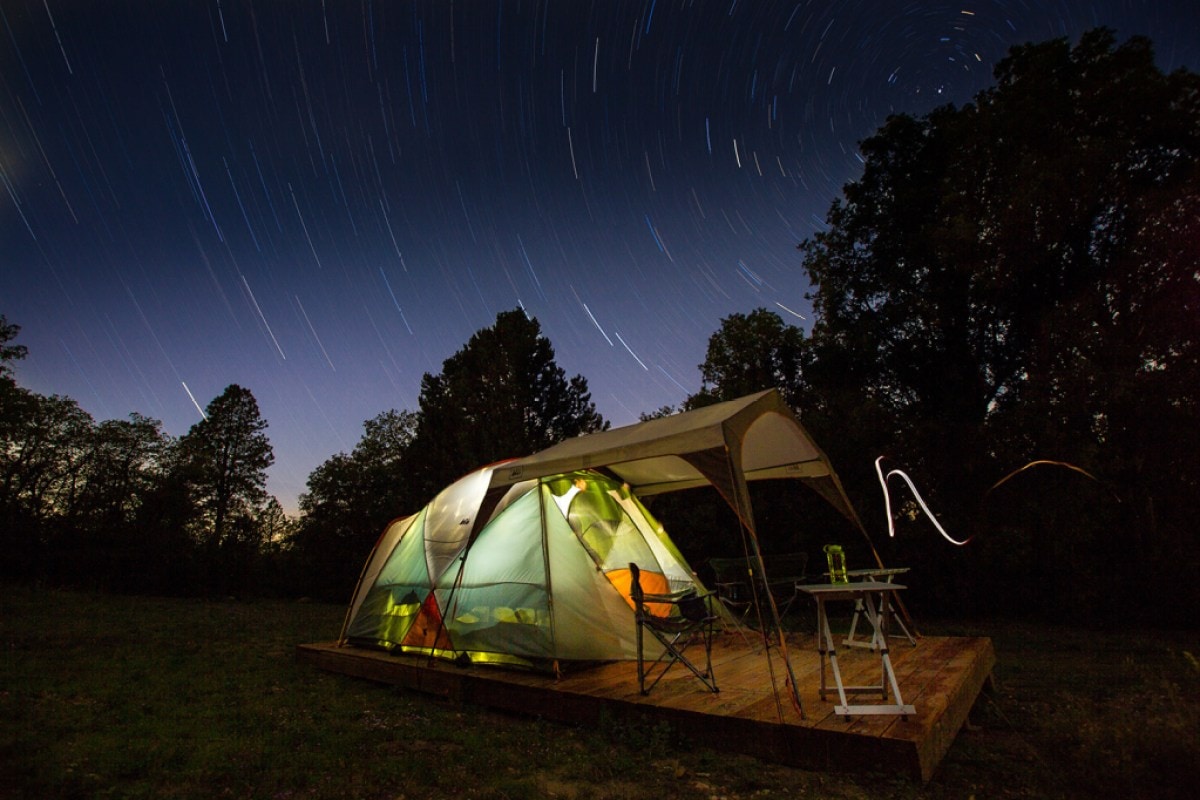 A camping tent is illuminated from within and the stars are photographed with a long exposure behind them