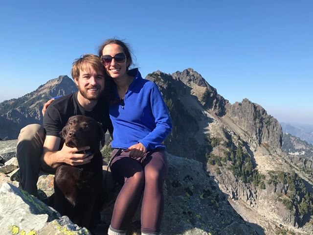A couple sits hugging their dog on the top of a summit, mountains in the background.