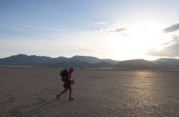 Thru hiker with pack on walks across cracked desert, mountain in the background.