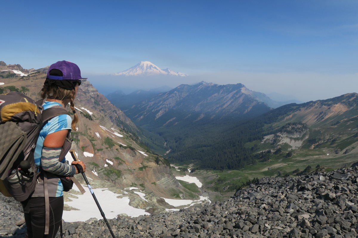 Thru hiker with back to camera looks out on a mountain view, blue skies above.