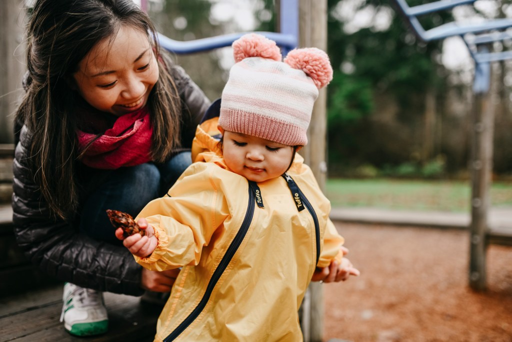 A woman supports a toddler in a yellow onesie as she plays on a playground