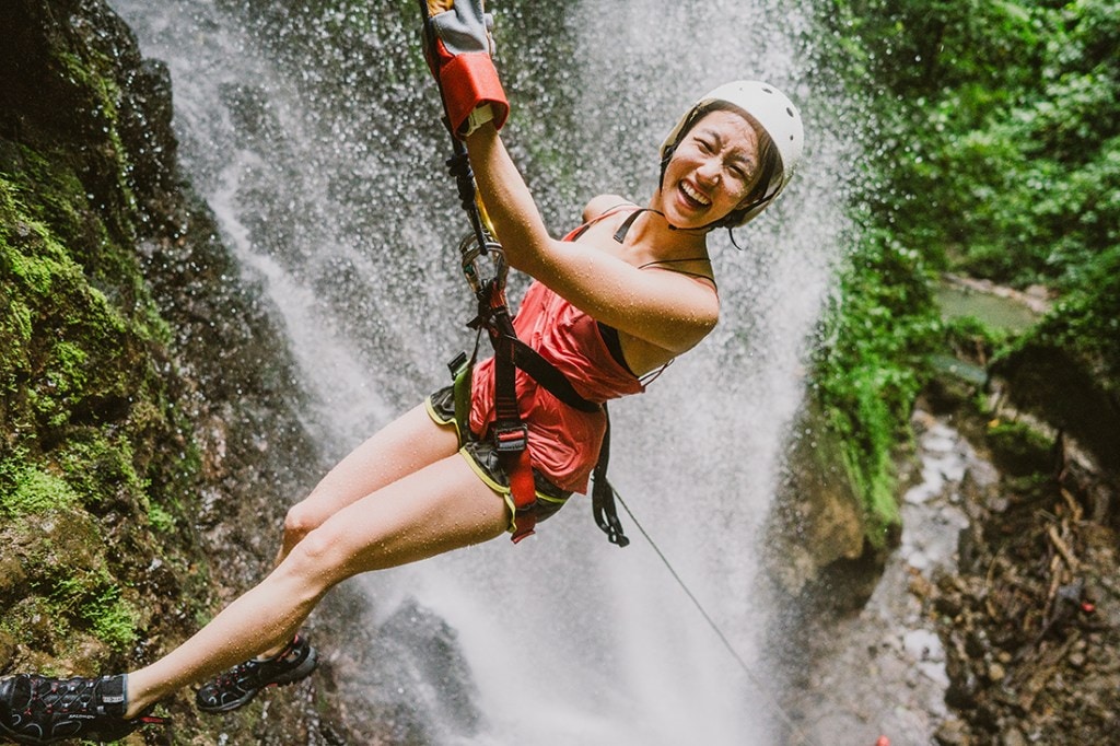 A woman is obviously stoked as she rappels in front of a lush, flowing waterfall. 