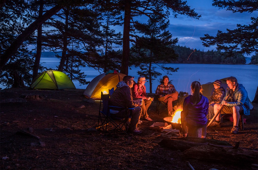An REI Adventures family trip enjoys the crackling firelight at their waterside campsite.