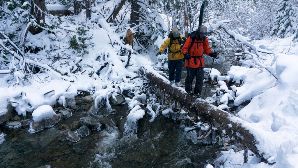 Danny Davis and Nick Russell walk with their snowboards across a snowy log over a creek.