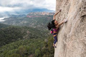 Olivia Hsu reaches dynamically for a hand hold on a steep rock cliff with a green valley stretching out below her.