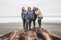 Four hikers stand close on top of a stump near the coast, with gray clouds in the distance.