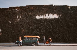 Three friends pack a green car with an orange surfboard on top. They are in front of a large ivy-covered wall.