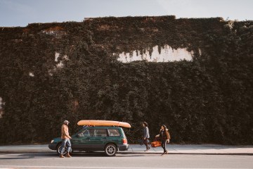 Three friends pack a green car with an orange surfboard on top. They are in front of a large ivy-covered wall.