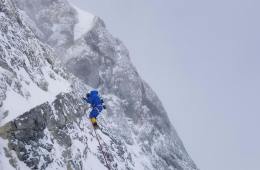 A climber in a down suit traverses across a steep rock face covered in patches of snow and ice with thick clouds in the background.