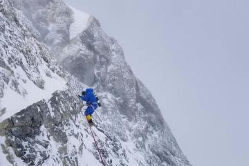 A climber in a down suit traverses across a steep rock face covered in patches of snow and ice with thick clouds in the background.