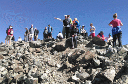Hikers stand along the top of a ridgeline, looking and pointing every which way under a bright-blue sky.