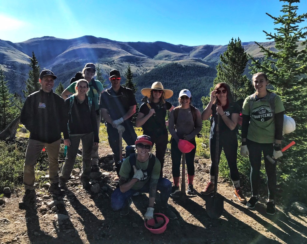 Volunteers with big smiles, heavy packs and mallets and helmets in their hands, standing along a ridgeline.