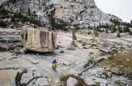 A lone rock climber walks down a low-angle slab of grey rock with a large boulder and ridge in the background.