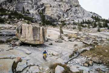 A lone rock climber walks down a low-angle slab of grey rock with a large boulder and ridge in the background.