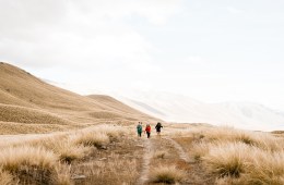 Three hikers in the distance walk toward the camera, a white sky above and dry grasses below.