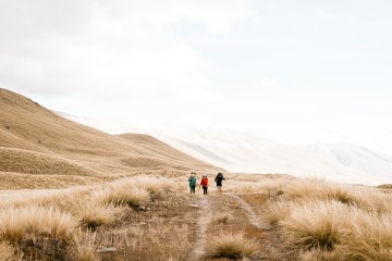 Three hikers in the distance walk toward the camera, a white sky above and dry grasses below.