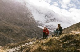 Two people sit on the ground, a huge snowy mountain is behind them.