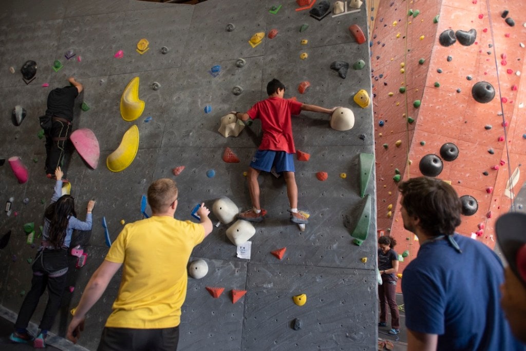 Swee Mana balances on while holds on an indoor bouldering wall while an onlooker points, giving instruction.
