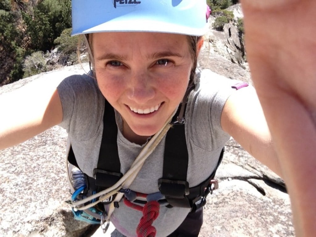 Beth Rodden, wearing a full-body climbing harness and helmet, takes a selfie on top of a rock dome.