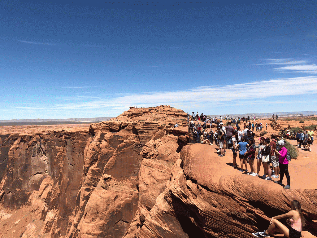 A crowd congregates on the side of a mesa to take in a sweeping view of the desert and the bright blue sky