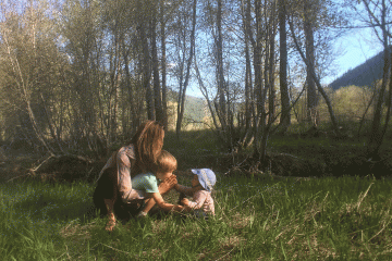 A woman and two small children practice yoga in a green field with sunlight shimmering in the background