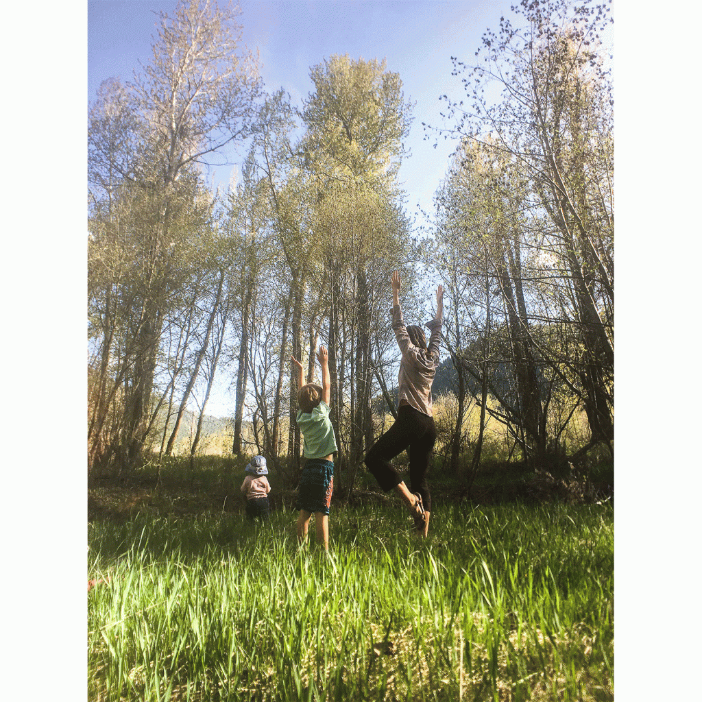 A woman and two young children practice the tree yoga pose in a green field with sunlight shimmering all around them