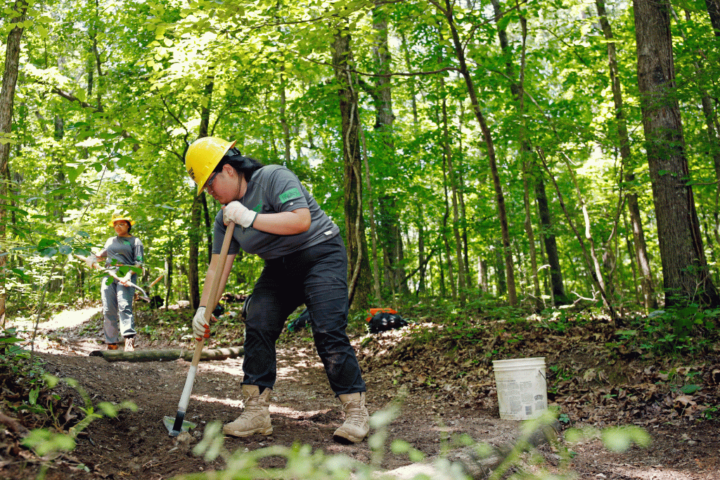 A woman maintains a trail using a shovel surrounded by lush forest