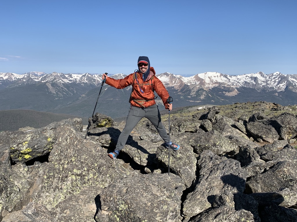 A man in a bright-orange jacket standing among mountains with a. blue sky behind him
