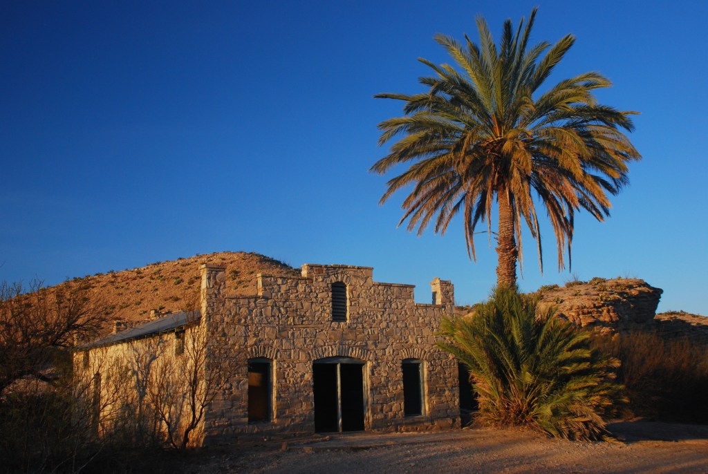 A palm tree stands next to the ruins of a post office as the sun sets over the landscape.