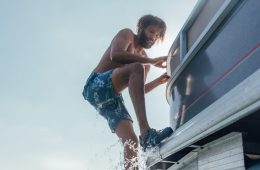 A man is climbing down off a boat to get into the water. He's wearing a bathing suit and KEEN sandals.