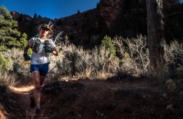 Clare Gallagher is running on a trail with mountains and blue sky behind her.