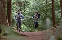A couple jogs through the forest together surrounded by tall evergreen trees
