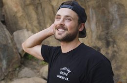 Dylan Efron smiles at the camera with one of his hands behind his head. He has on a backwards black baseball cap and there's a boulder behind him.
