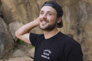Dylan Efron smiles at the camera with one of his hands behind his head. He has on a backwards black baseball cap and there's a boulder behind him.