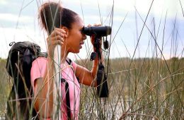 Corina Newsome stands with her profile to the camera, holding binoculars up to her face while birdwatching. She's wearing a pink shirt and has on a black backpack.