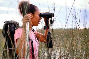 Corina Newsome stands with her profile to the camera, holding binoculars up to her face while birdwatching. She's wearing a pink shirt and has on a black backpack.