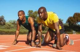 David Brown is kneeling down on the track in a blue tank top next to his running guide, Jerome Avery who is tied to him with a thread between their hands. Jerome is also kneeling down next to Davis with a yellow t-shirt on.