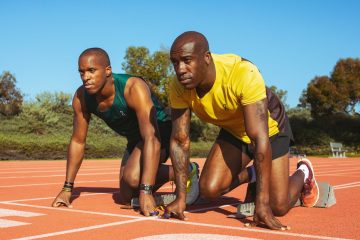 David Brown is kneeling down on the track in a blue tank top next to his running guide, Jerome Avery who is tied to him with a thread between their hands. Jerome is also kneeling down next to Davis with a yellow t-shirt on.
