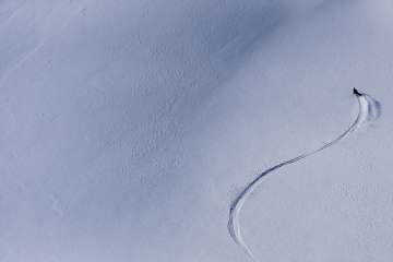 Michelle Parker captured from an aerial camera skiing down a mountain. She is a tiny bright spot in a sea of white.