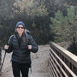 Woman hiker wearing sunglasses and using trekking poles as she crosses a wooden boardwalk bridge