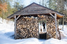 Joseph Mulherin, of nothing.nowhere, is silhouetted by a shed filled with chopped wood as he swings an axe over a piece of wood.
