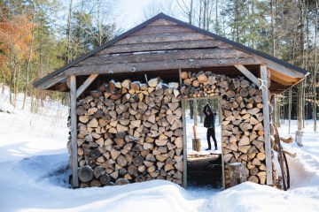 Joseph Mulherin, of nothing.nowhere, is silhouetted by a shed filled with chopped wood as he swings an axe over a piece of wood.