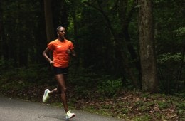 Sika Henry is running on a gravel path in an orange shirt and black shorts with trees in the background.