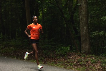 Sika Henry is running on a gravel path in an orange shirt and black shorts with trees in the background.