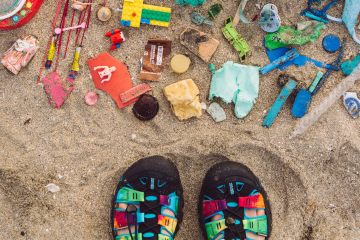A pair of bright, rainbow KEEN shoes next to a pile of brightly colored objects on a beach.