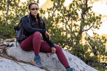 Nicole Snell, wearing purple pants, hiking boots and a black jacket, is sitting on a rock and looking at the camera with trees in the background.