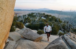 Lesford Duncan is running up a trail with a white HOKA shirt on and there is a mountain range in the background.