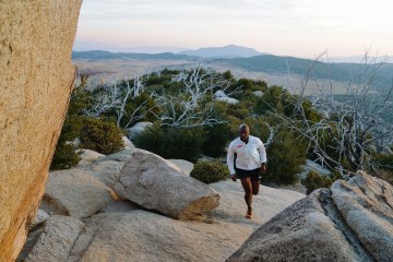 Lesford Duncan is running up a trail with a white HOKA shirt on and there is a mountain range in the background.