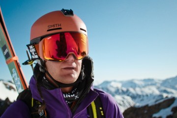 Connor Ryan looks at the camera with ski goggles on and skis strapped to his back. In the background is a mountain range.