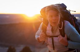 Erin Parisi is looking at the camera with the sun behind her and a backpack on her back. There's a mountain range in the distance.